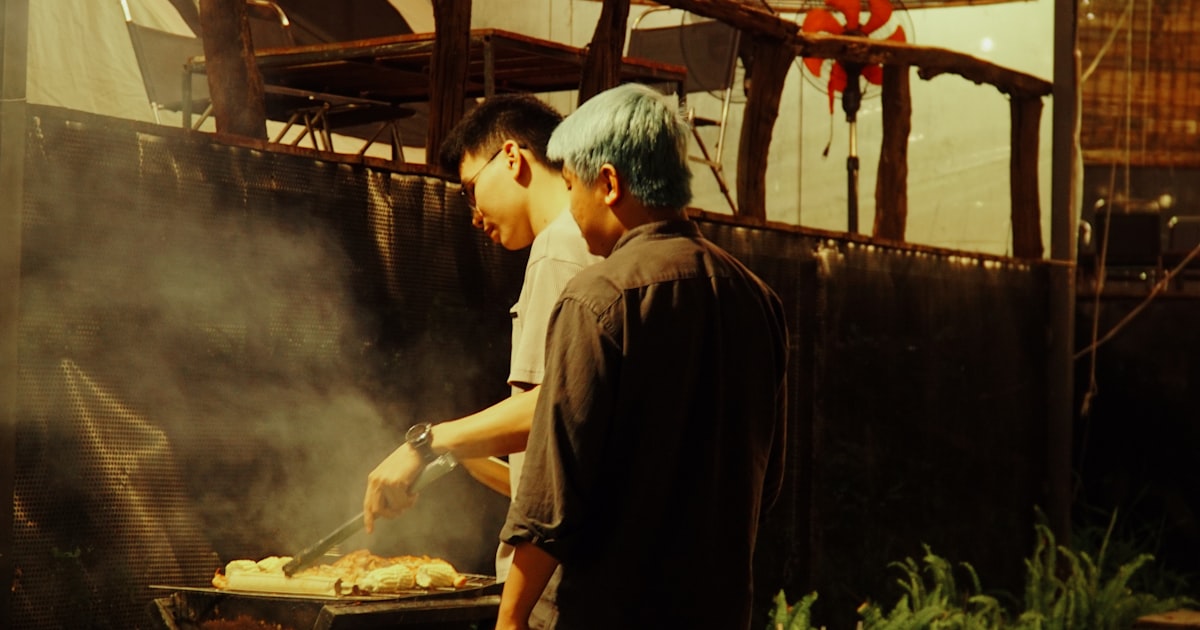 Two men grilling food outdoors at night. — Clube Prime, Empório Família Rodrigues